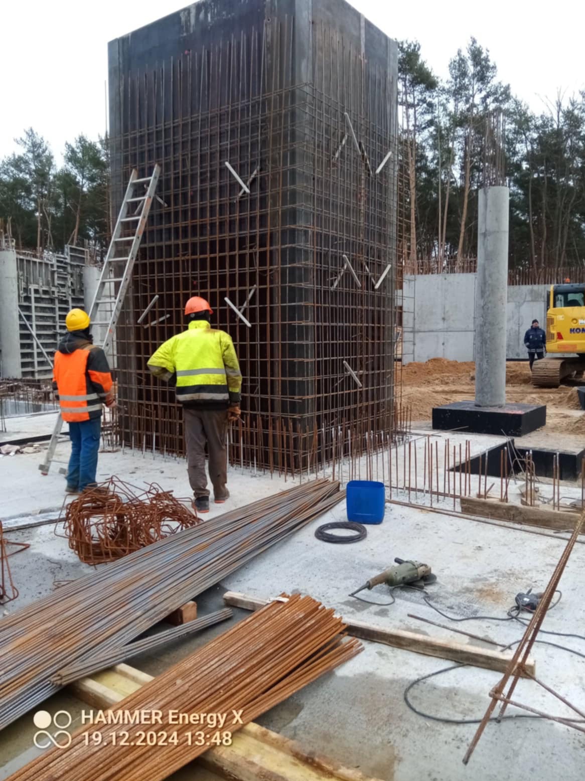 Wiązanie zbrojenia pionowego słupa z wykorzystaniem drabiny, na budowie widoczny wykop i maszyna budowlana.