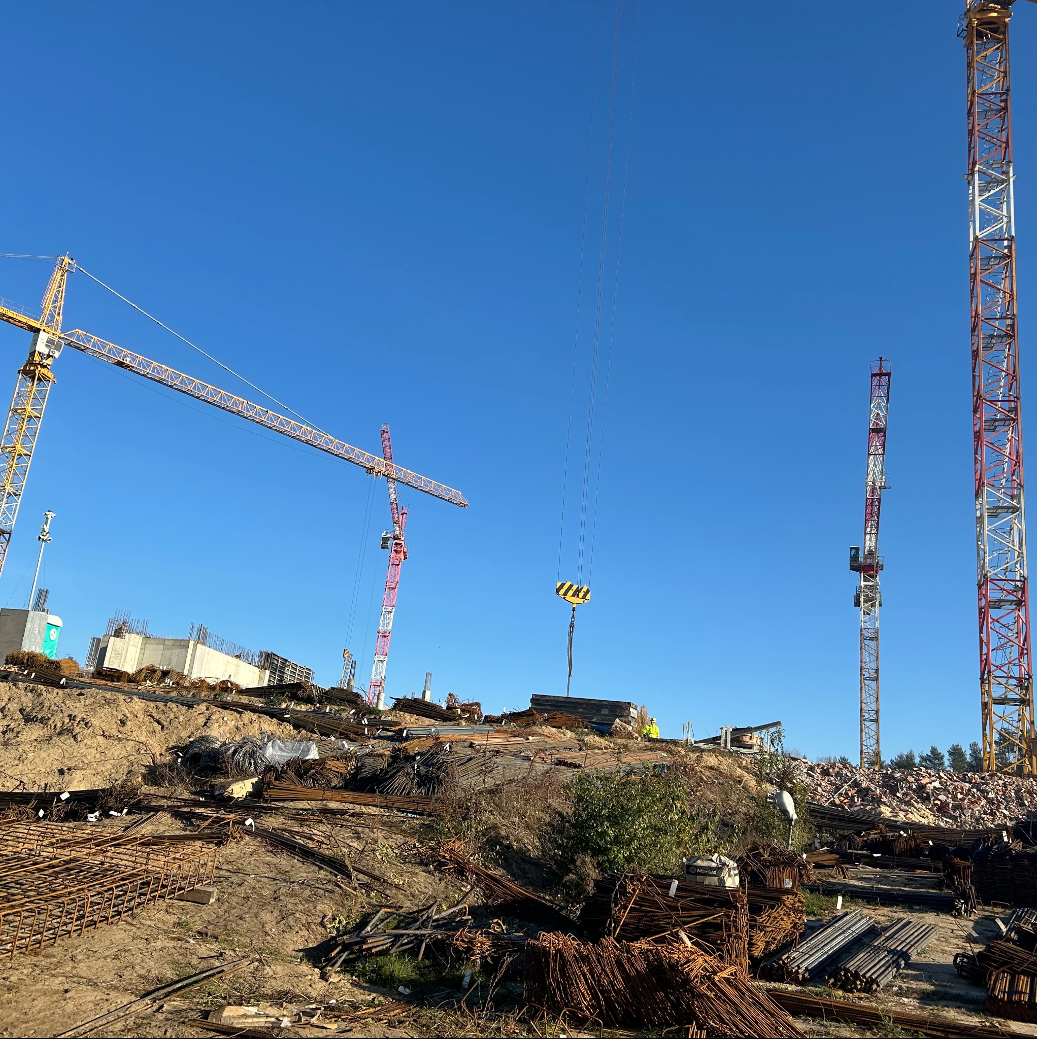 Plac budowy z trzema żurawiami wieżowymi, zbrojeniami stalowymi i wylewanymi elementami konstrukcji żelbetowej w tle, widok w słoneczny dzień.