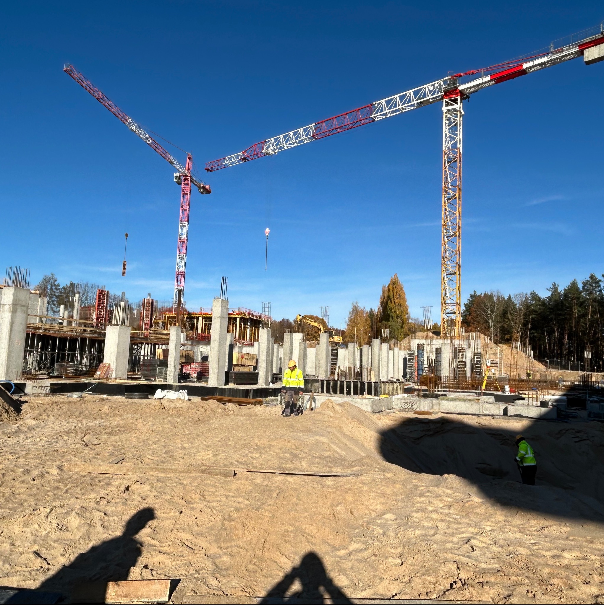 Budowa budynku żelbetowego w trakcie realizacji, widoczne szalunki, zbrojenia, betonowe słupy, dwóch pracowników w odblaskowych kamizelkach oraz dwa żurawie wieżowe na tle niebieskiego nieba i drzew.