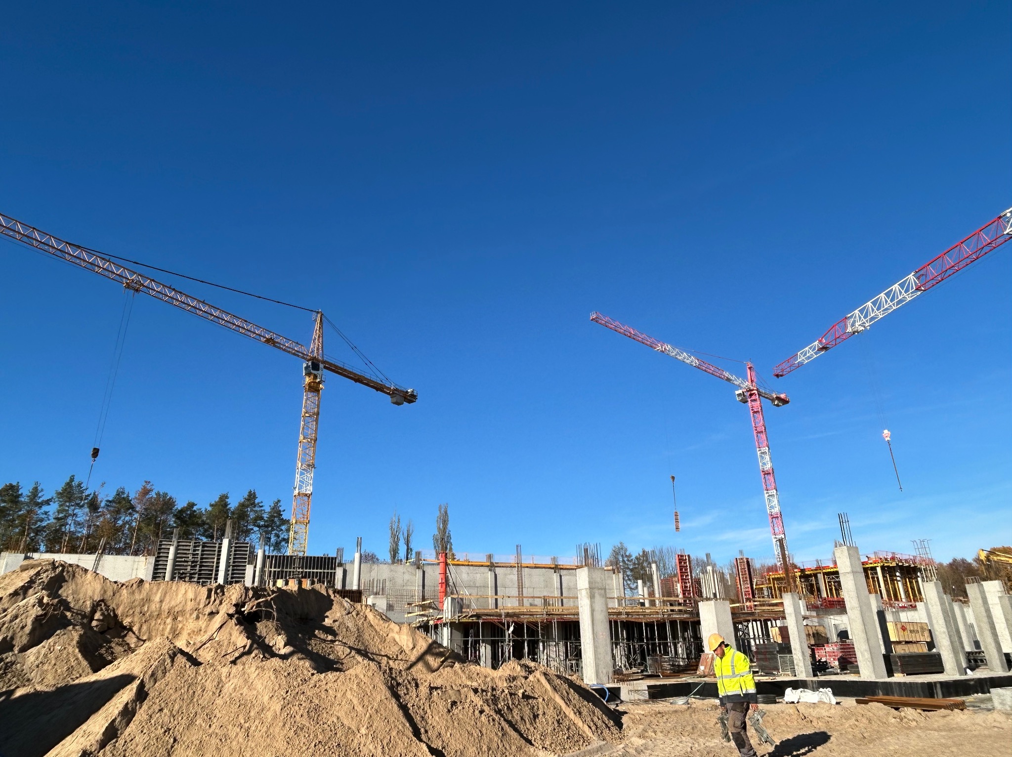 Budowa budynku żelbetowego w słoneczny dzień, widoczne trzy żurawie budowlane, sterty piasku i pracownik w żółtej kamizelce.