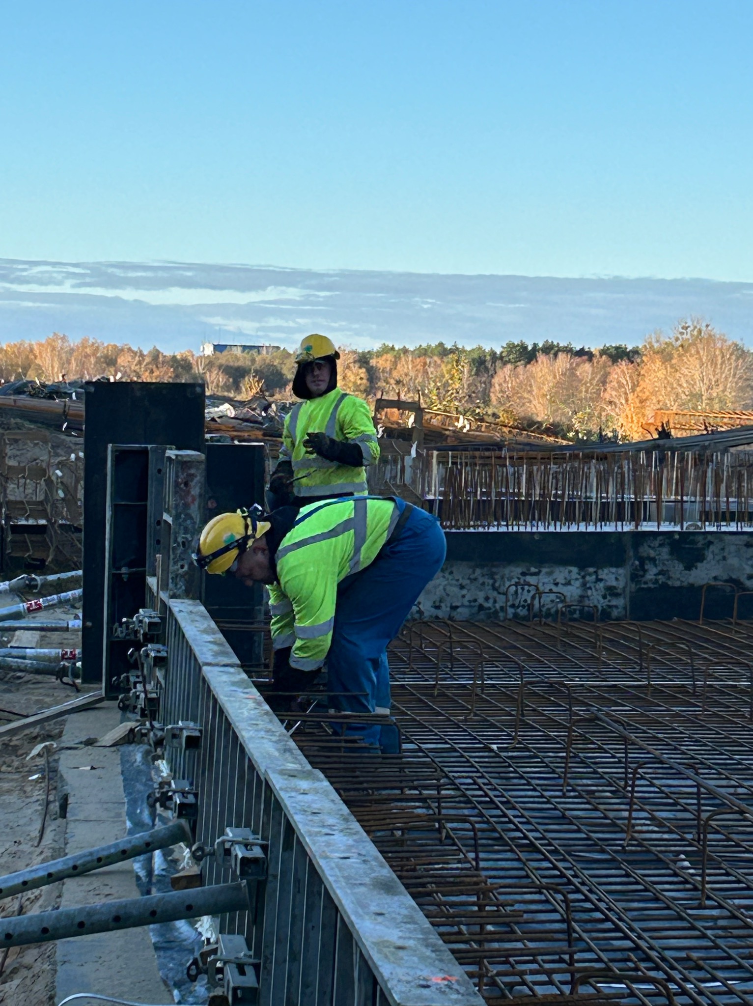 Dwóch pracowników w żółtych kamizelkach i kaskach montuje zbrojenie na budowie, widoczne szalunki i stalowe pręty, w tle drzewa i niebo.