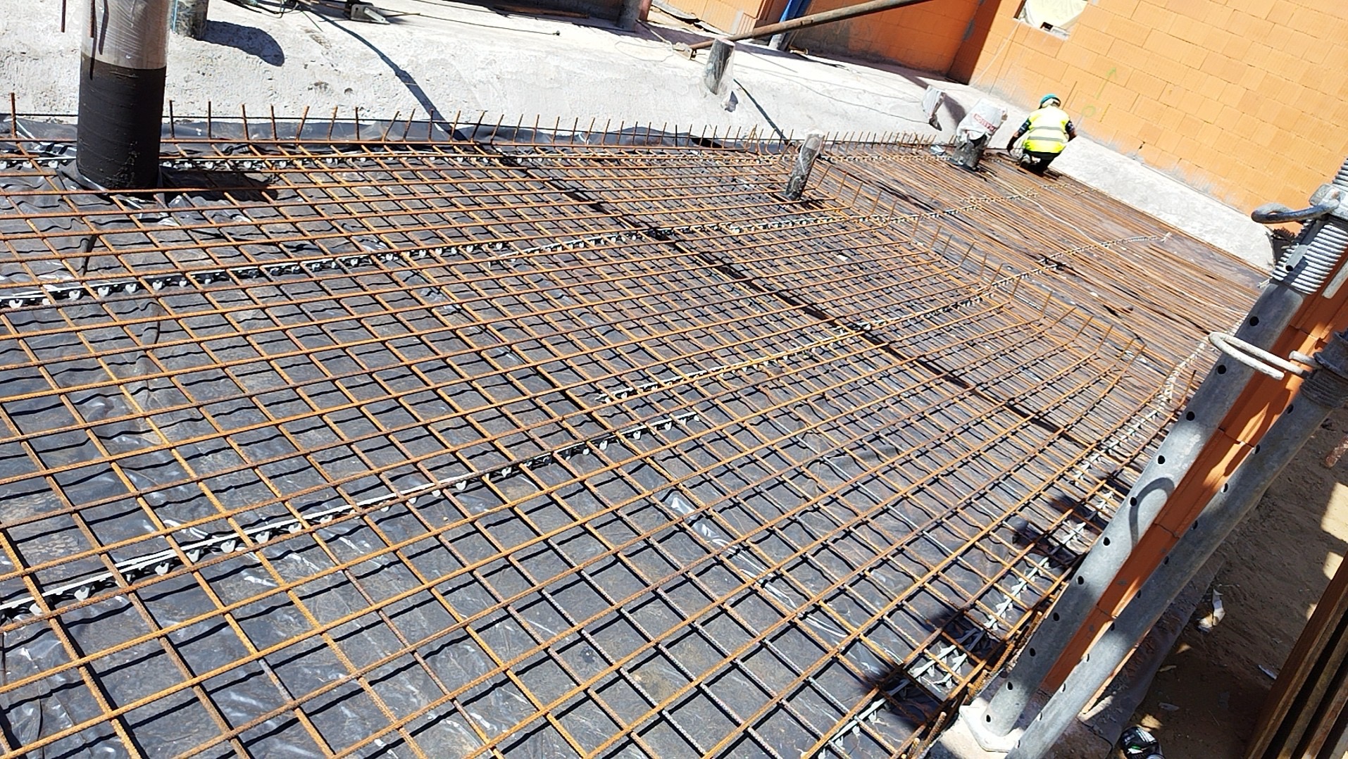 Siatka zbrojeniowa ułożona na czarnej folii, widoczny pracownik w żółtej kamizelce i kasku podczas montażu, rury wentylacyjne i ceglany budynek w tle.