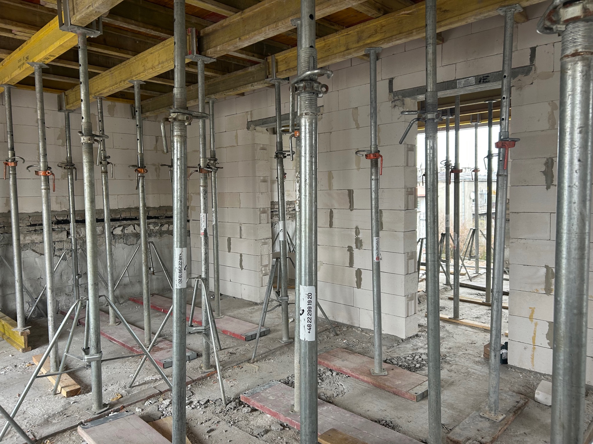 Wnętrze budynku w trakcie budowy z białych bloczków, podparte metalowymi stemplami i żółtymi belkami stropowymi. Widoczne otwory okienne i drzwiowe.