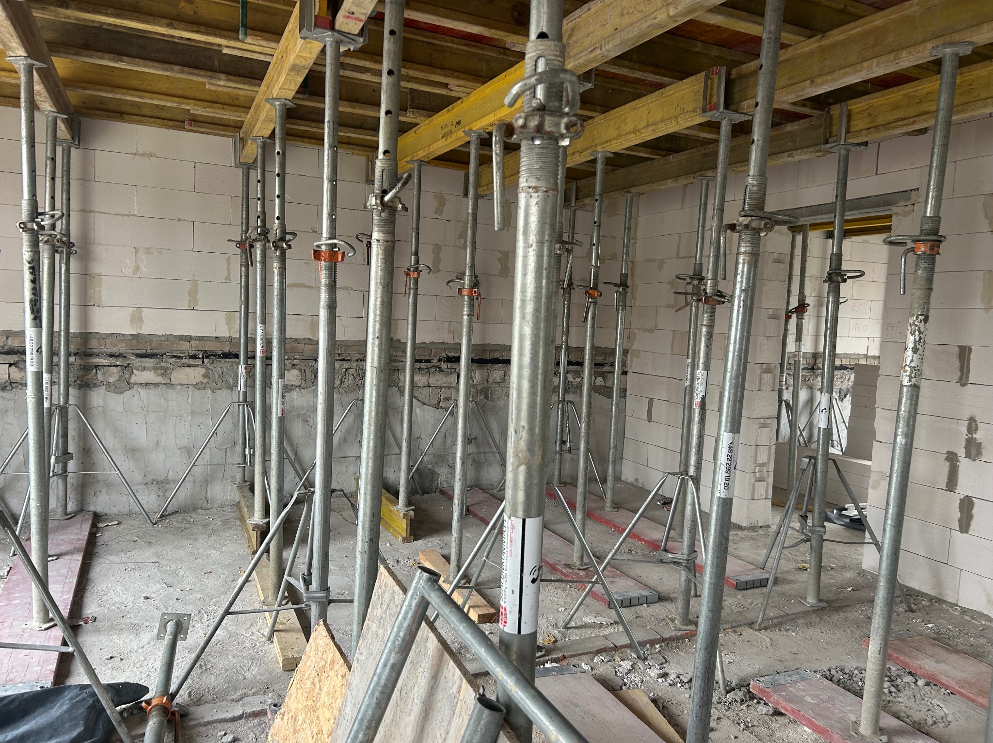 Wnętrze budynku w trakcie budowy z widocznymi podporami stropowymi i elementami szalunku. Ściany z bloczków, surowy stan wykończenia.