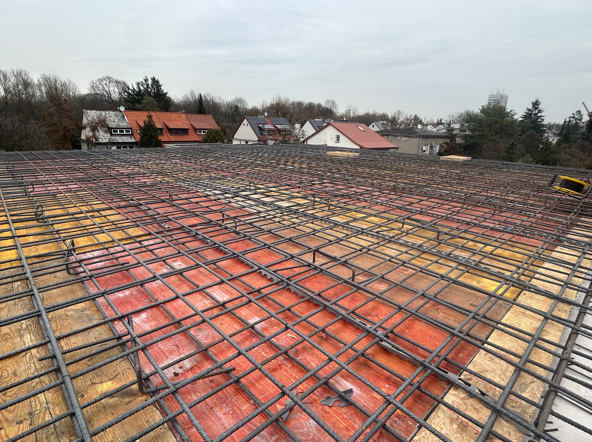 Zbrojenie stropu na deskowaniu z widokiem na zabudowę jednorodzinną w tle. Siatka z prętów stalowych tworzy regularną strukturę. Widoczne elementy deskowania.