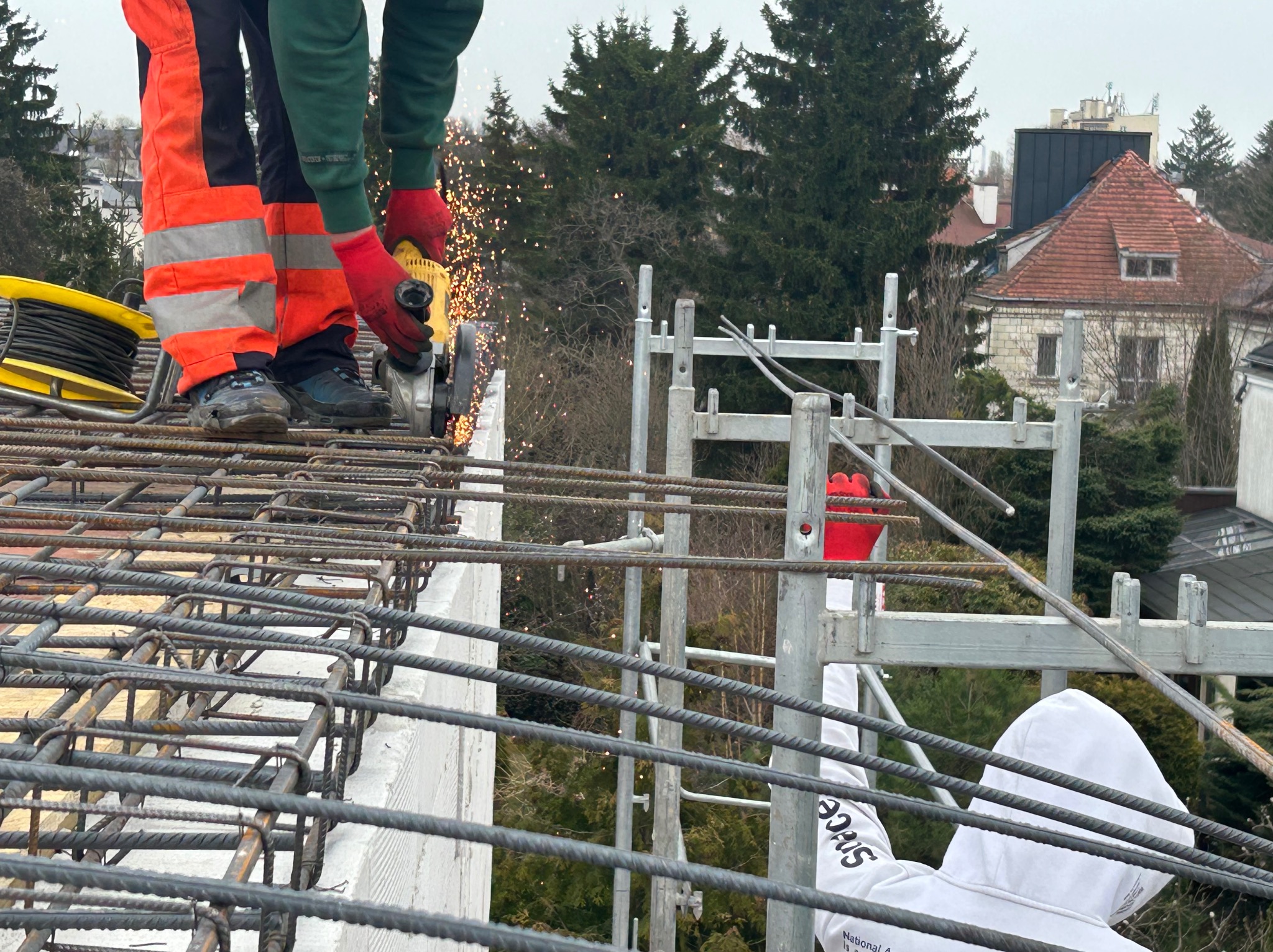 Praca zbrojarska w Klukówku: zbrojarz w odzieży roboczej tnie pręt zbrojeniowy szlifierką kątową na tle konstrukcji zbrojeniowej i zieleni.