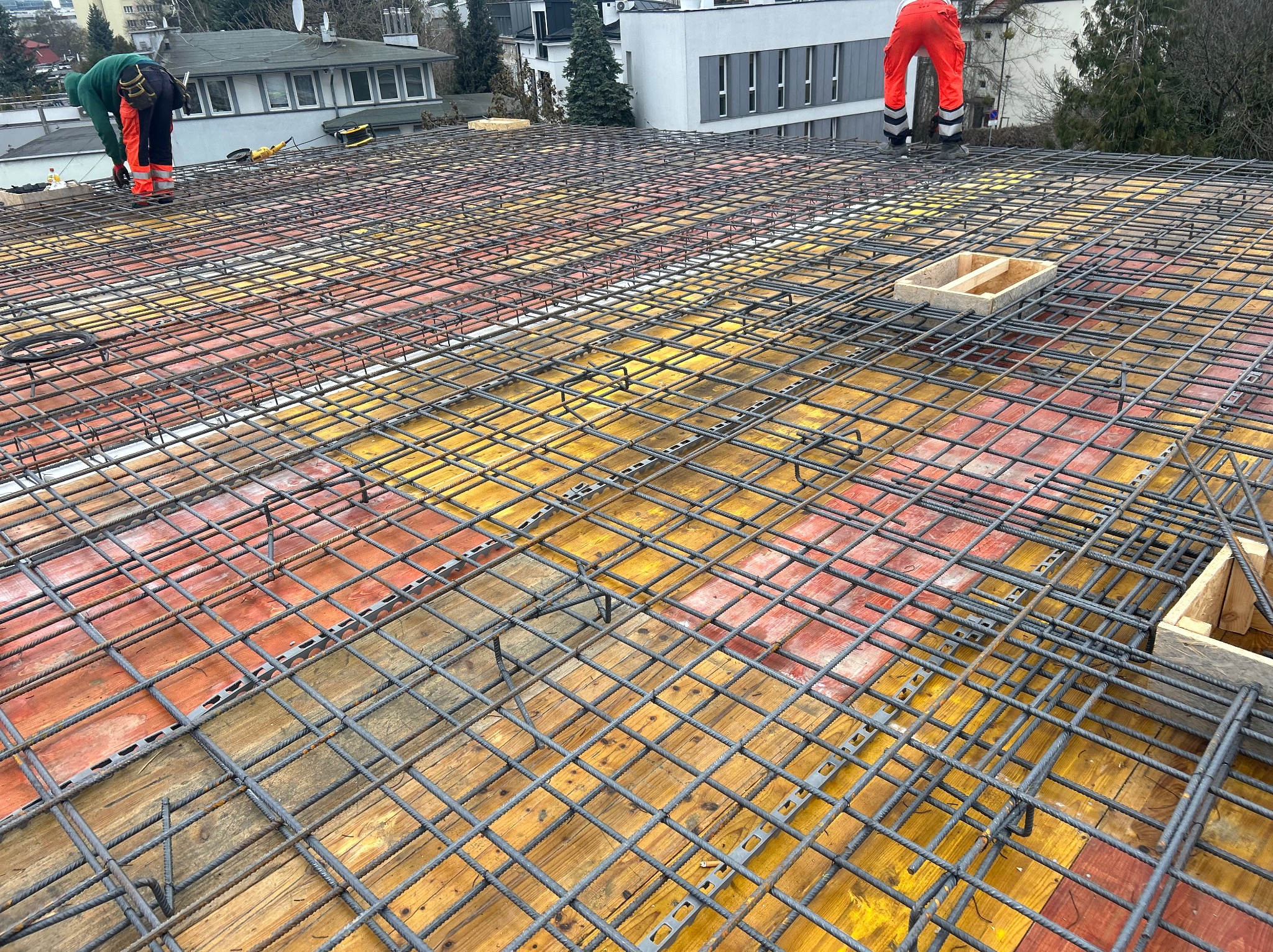 Siatka zbrojeniowa na deskowaniu, przygotowana do zalania betonem. W tle pracownicy w odzieży roboczej. Widok z góry, szeroki kadr.