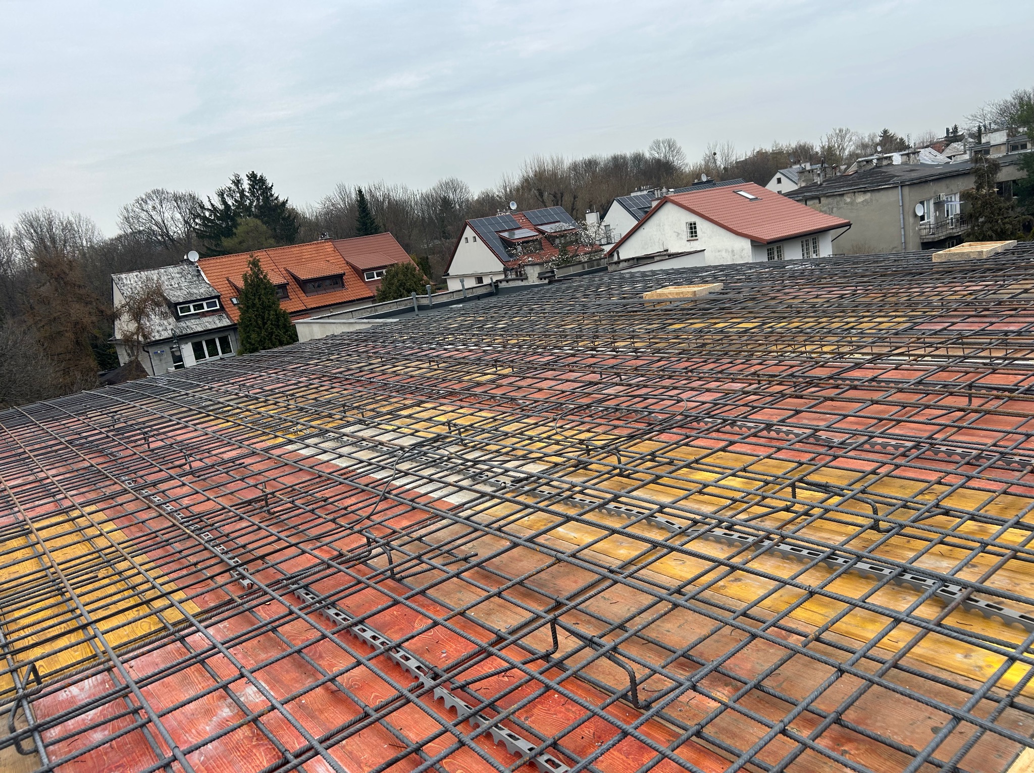 Siatka zbrojeniowa na deskowaniu, widok z góry na tle zabudowy jednorodzinnej. Elementy dystansowe pod zbrojeniem. Szara kolorystyka nieba.