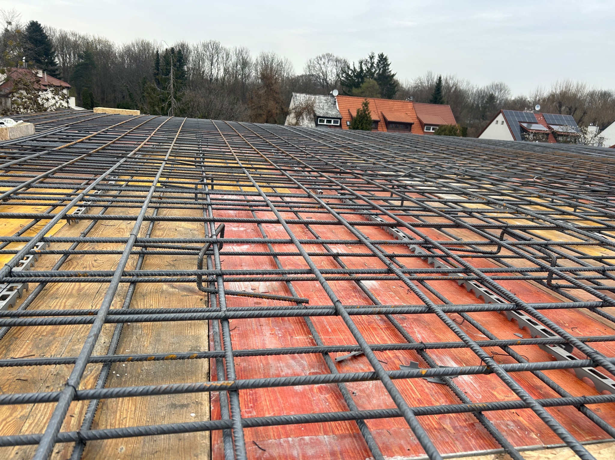 Siatka zbrojeniowa na deskowaniu, widok z góry. W tle zabudowa jednorodzinna i drzewa. Zbrojenie ułożone równomiernie, przygotowane do zalania betonem.