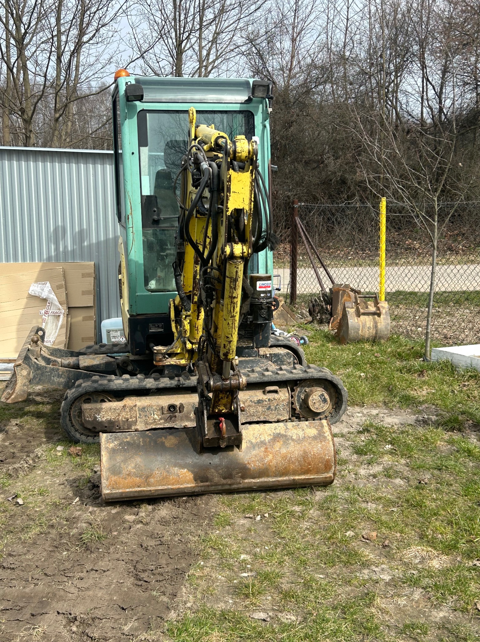 Żółto-zielona mini koparka z łyżką na trawiastym terenie, widok z przodu, w tle metalowe ogrodzenie i drzewa bez liści. Na pierwszym planie ślady opon na ziemi.