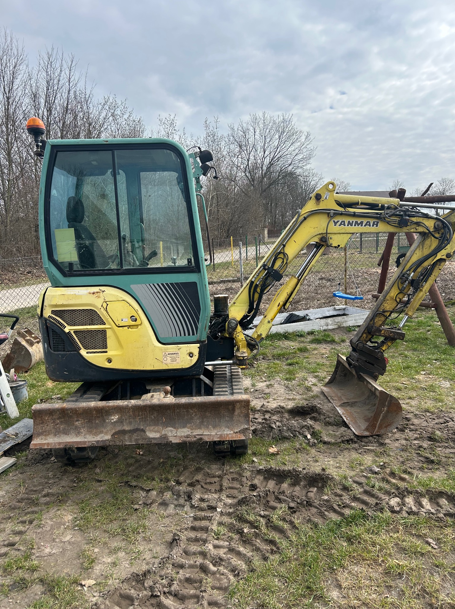 Żółto-zielona mini koparka Yanmar na trawiastym terenie z widocznymi śladami gąsienic, w tle drzewa i ogrodzenie z siatki.