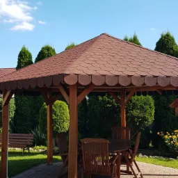 Drewniana altana ogrodowa z brązowym dachem krytym gontem bitumicznym, wyposażona w stół i krzesła na brukowanym patio, otoczona zielenią.