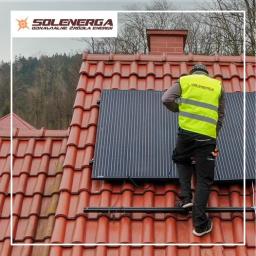 Solenerga - Pracownik w żółtej kamizelce zakłada panel słoneczny na dachu z czerwonej dachówki, widoczny komin i las w tle.