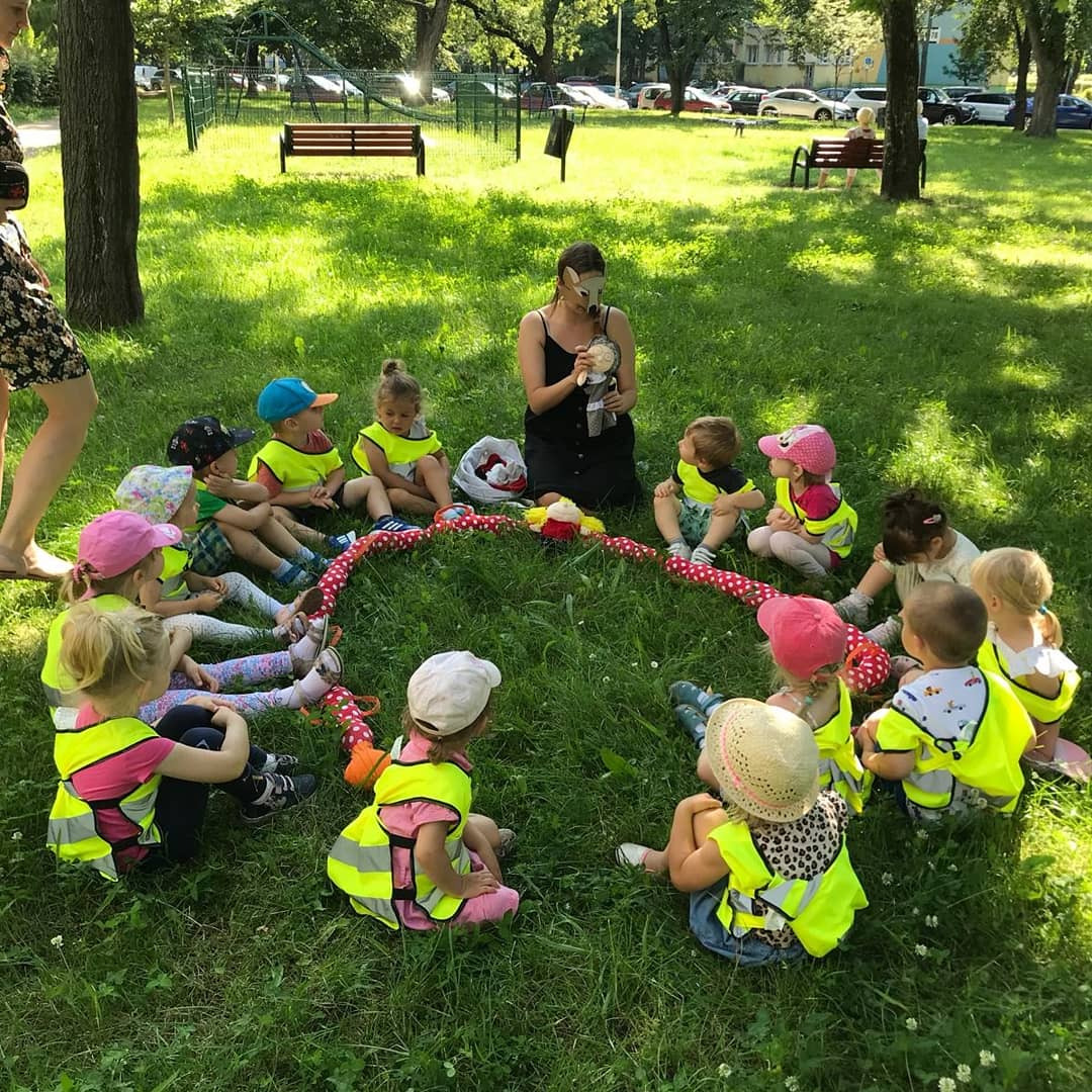 Grupa dzieci w kamizelkach odblaskowych siedzi na trawie w parku, otoczona czerwoną, wzorzystą liną; opiekunka w masce lisa opowiada historię, trzymając lalkę.