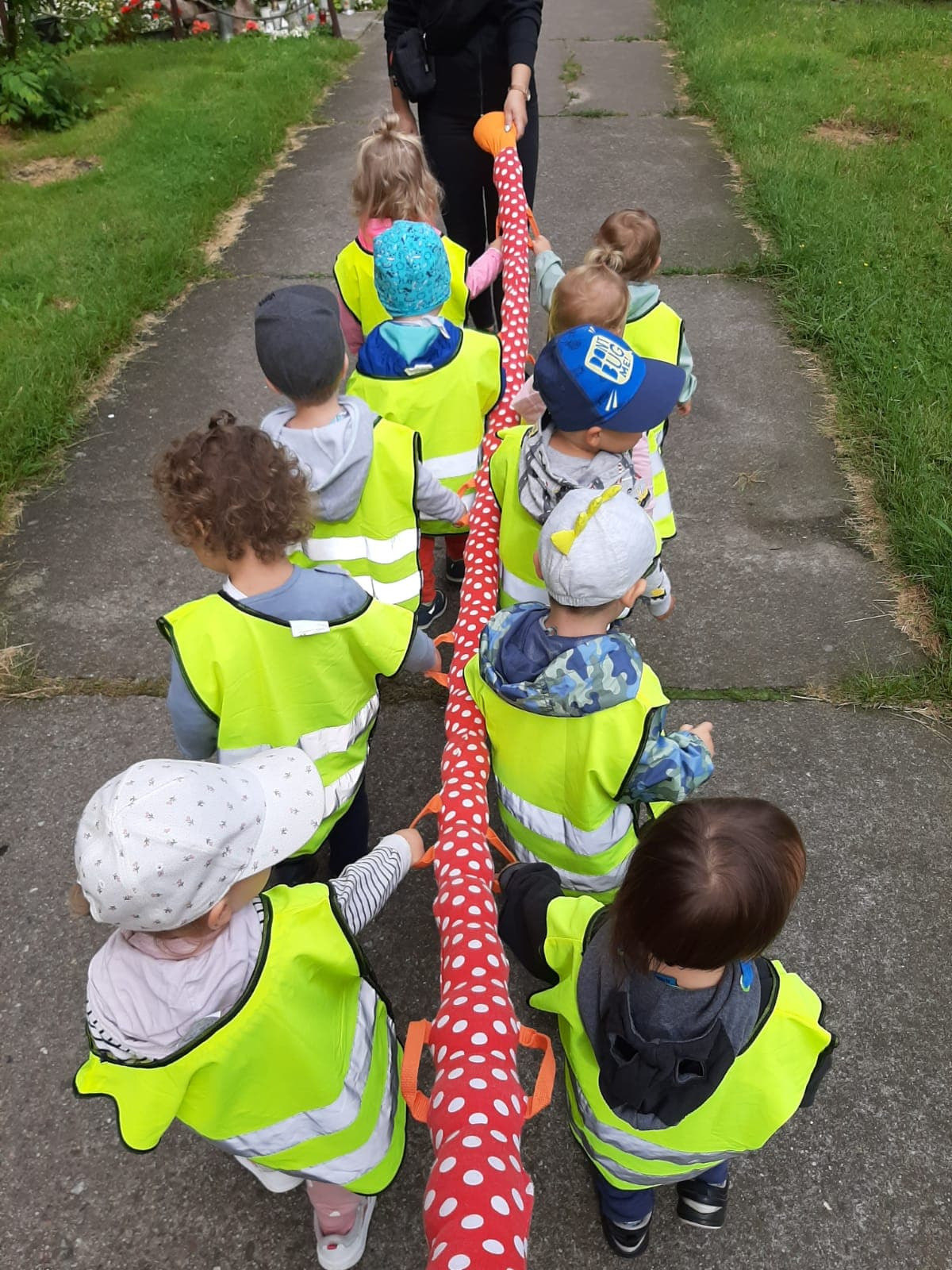 Grupa dzieci w jaskrawych kamizelkach idzie ścieżką trzymając długą, czerwoną zabawkę w białe kropki, przypominającą węża lub gąsienicę.