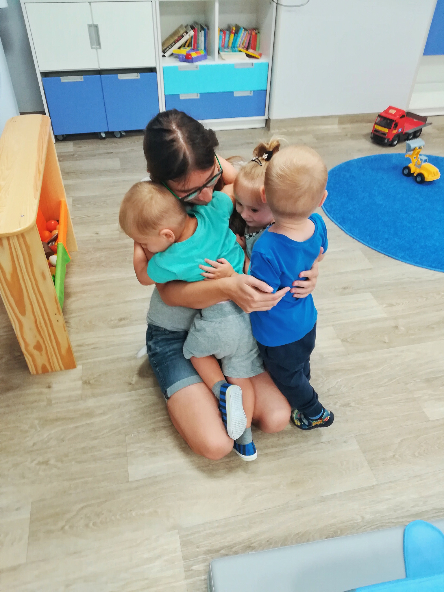 Opiekunka w okularach przytula troje dzieci w sali zabaw z drewnianą podłogą, w tle regał z zabawkami i niebieski dywan z zabawkami.