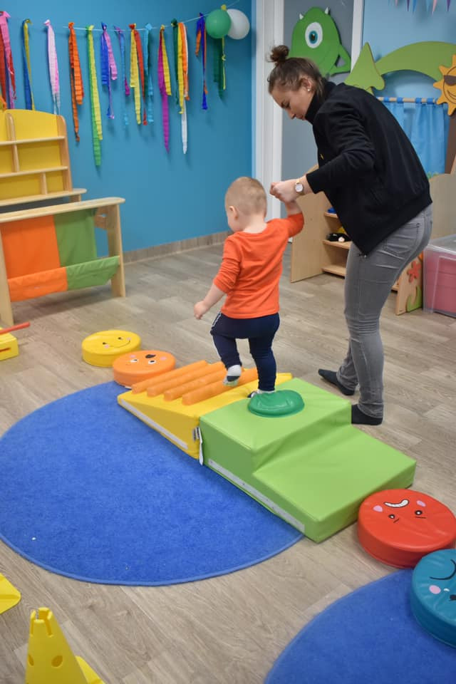 Chłopiec w pomarańczowej koszulce ćwiczy motorykę na kolorowym torze przeszkód w żłobku, trzymany za rękę przez opiekunkę.