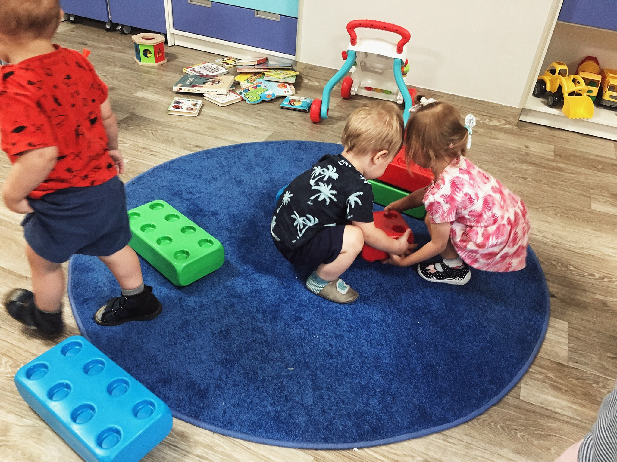 Dwoje dzieci bawi się klockami na okrągłym, niebieskim dywanie w pokoju zabaw, w tle widoczne półki z zabawkami i książkami.