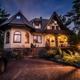 Elegancki pensjonat z podświetlonym wejściem, widoczny fragment samochodu na brukowanym podjeździe, ujęcie wieczorne.