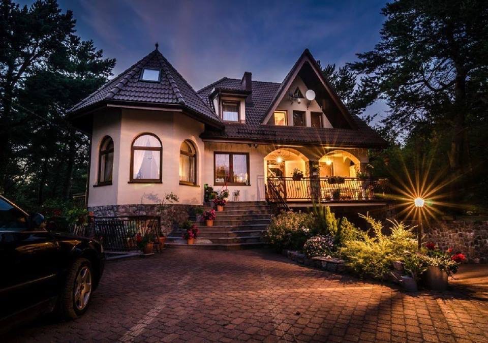 Elegancki pensjonat z podświetlonym wejściem, widoczny fragment samochodu na brukowanym podjeździe, ujęcie wieczorne.