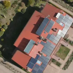 Widok z drona na duży budynek z czerwoną dachówką, na której zamontowane są panele słoneczne w różnych konfiguracjach; otoczenie budynku to zadbany ogród z geometrycznymi rabatami i ścieżkami.