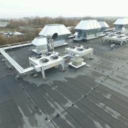 Klimatyzacja.in - Instalacja wentylacyjna z centralami na dachu budynku w Warszawie, widoczne liczne przewody i wentylatory, zarys miasta w tle.