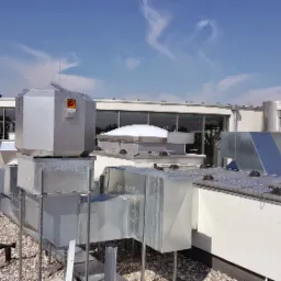 Instalacja wentylacyjna na dachu budynku z widocznymi kanałami wentylacyjnymi, centralą wentylacyjną z przyciskiem awaryjnym i elementami rekuperacji, na tle błękitnego nieba z delikatnymi obłokami.