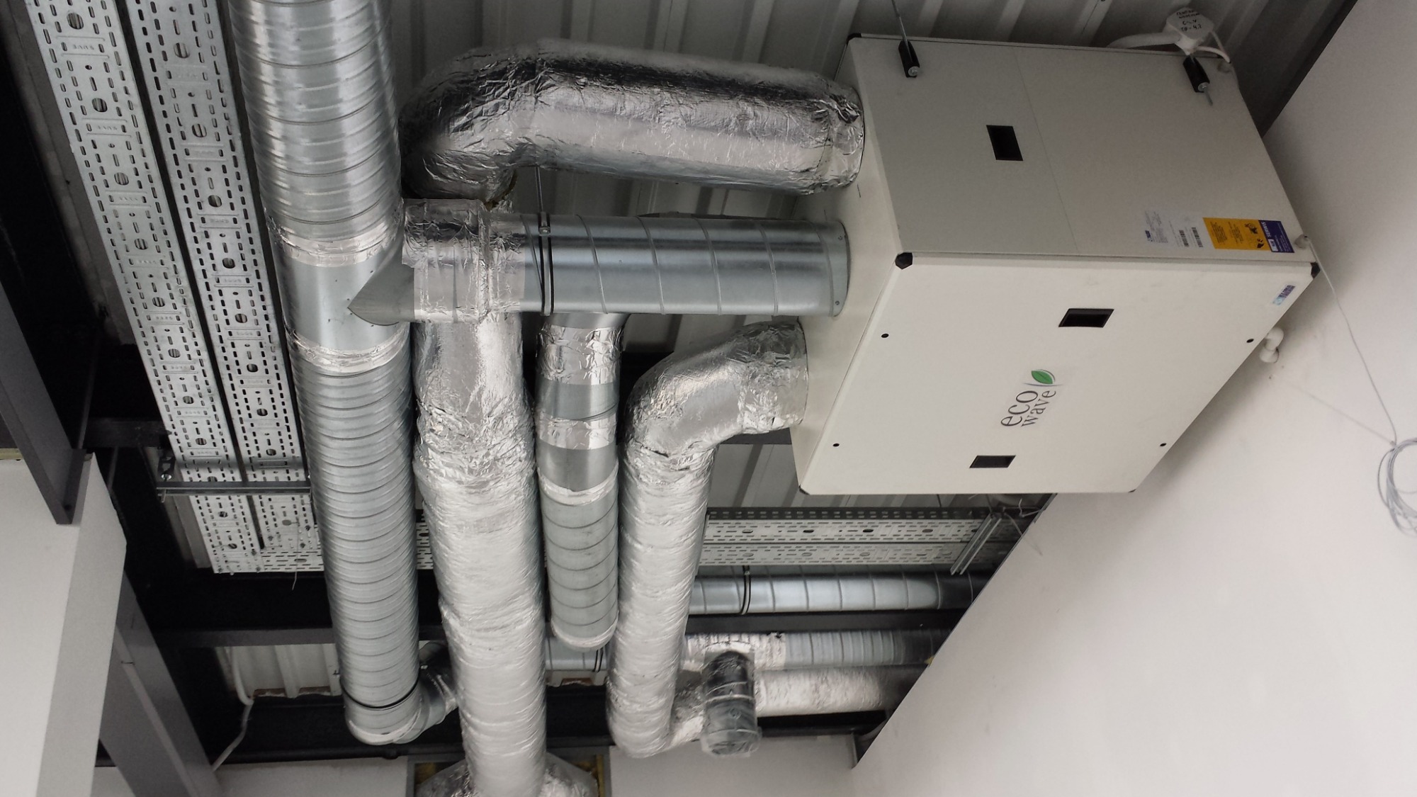 Zamontowany system z centralą wentylacyjną Eco Wave, rury wentylacyjne izolowane folią aluminiową i korytka kablowe pod sufitem w budynku komercyjnym.