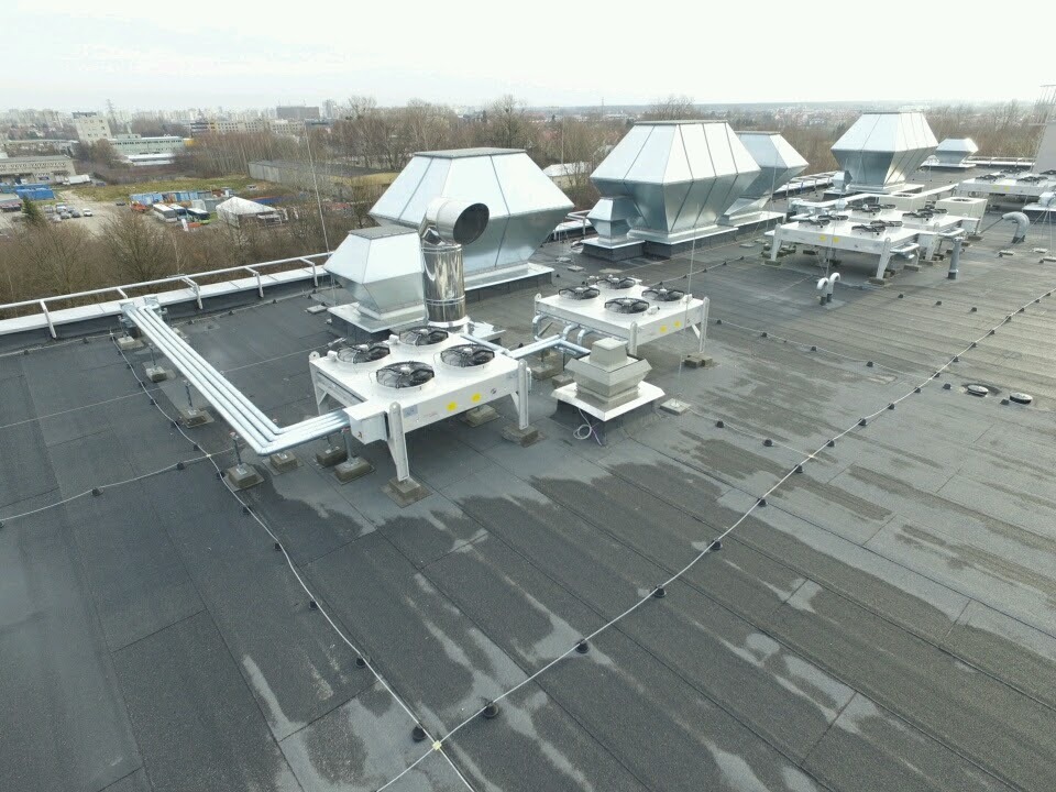 Instalacja wentylacyjna z centralami na dachu budynku w Warszawie, widoczne liczne przewody i wentylatory, zarys miasta w tle.