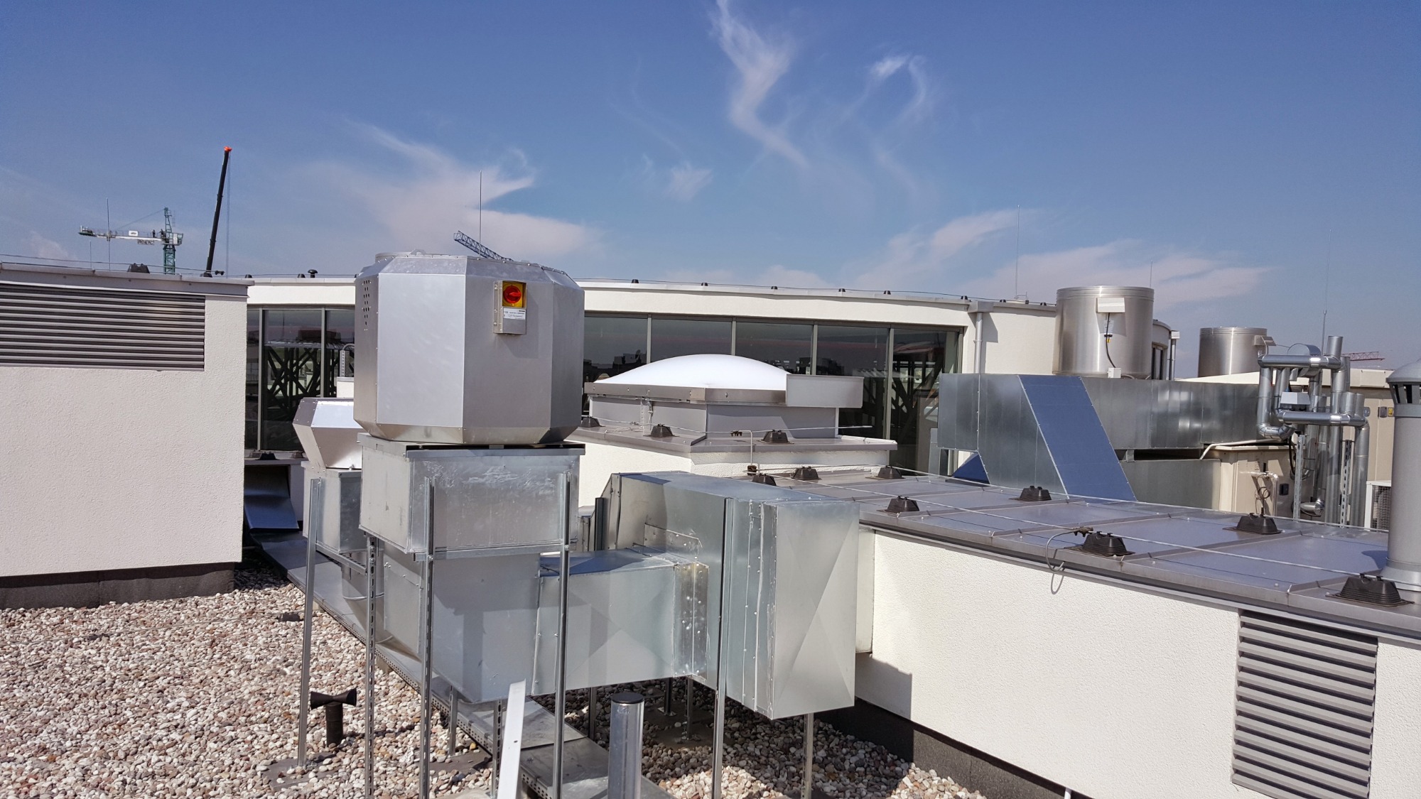 Instalacja wentylacyjna na dachu budynku z widocznymi kanałami wentylacyjnymi, centralą wentylacyjną z przyciskiem awaryjnym i elementami rekuperacji, na tle błękitnego nieba z delikatnymi obłokami.