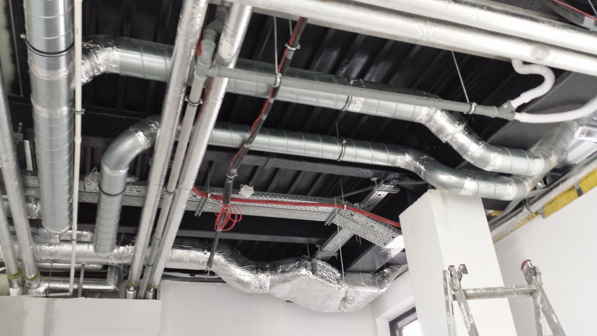 Widok instalacji wentylacyjnej pod sufitem, z widocznymi rurami aluminiowymi, przewodami elektrycznymi i drabinką opartą o białą kolumnę.