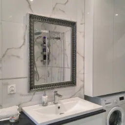 Jasna łazienka z umywalką wbudowaną w czarny blat, lustrem w ozdobnej ramie i pralką Beko w tle, płytki imitujące marmur na ścianie.