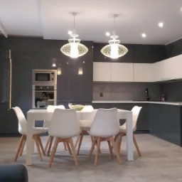 Nowoczesna kuchnia z szarymi szafkami i białymi górnymi szafkami, oświetlona dwoma dekoracyjnymi lampami wiszącymi nad białym stołem z krzesłami.