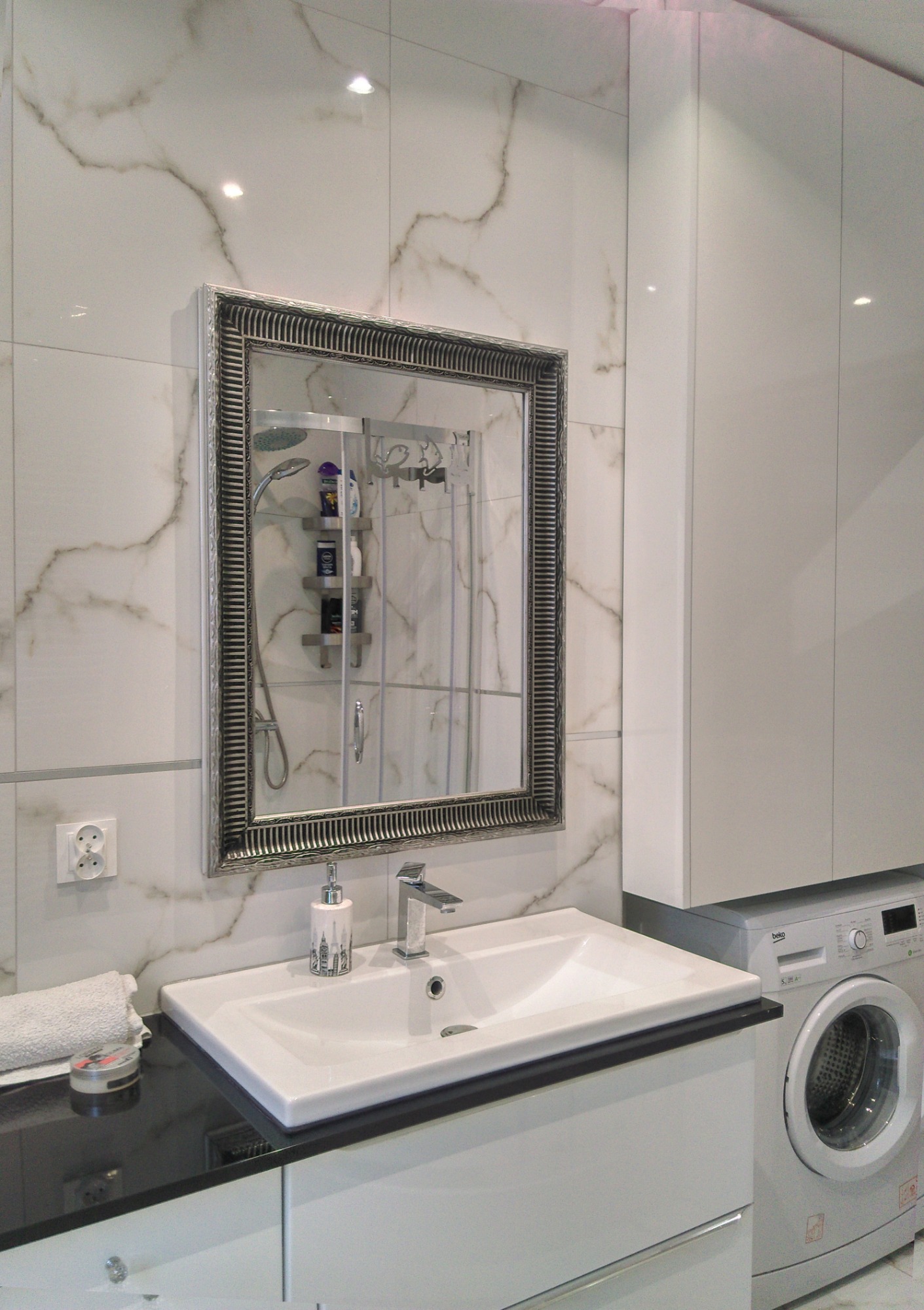 Jasna łazienka z umywalką wbudowaną w czarny blat, lustrem w ozdobnej ramie i pralką Beko w tle, płytki imitujące marmur na ścianie.