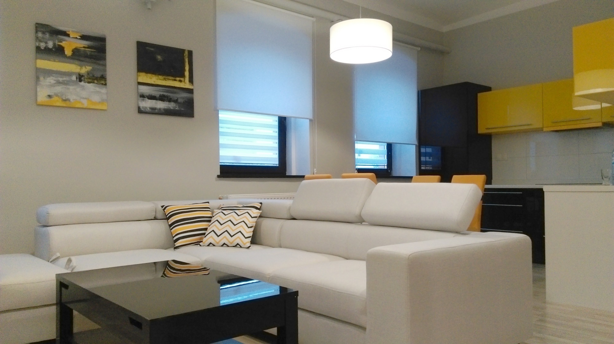 Jasny salon z białym narożnikiem, czarnym stolikiem kawowym i żółtymi akcentami w postaci obrazów i szafek kuchennych, oświetlony lampą wiszącą.