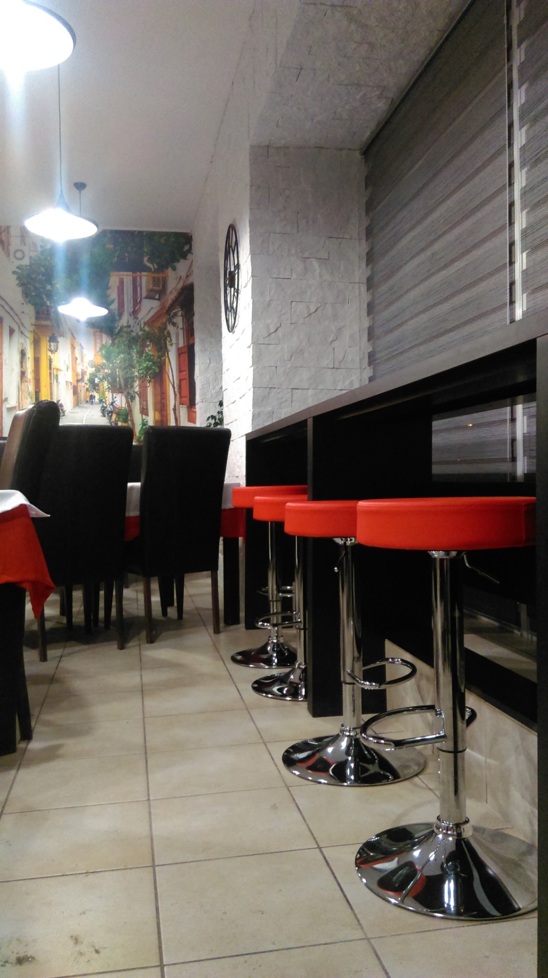 Wnętrze lokalu gastronomicznego z wysokimi, czerwonymi krzesłami barowymi przy czarnym blacie, w tle krzesła z ciemnymi obiciami i dekoracyjna fototapeta uliczki.