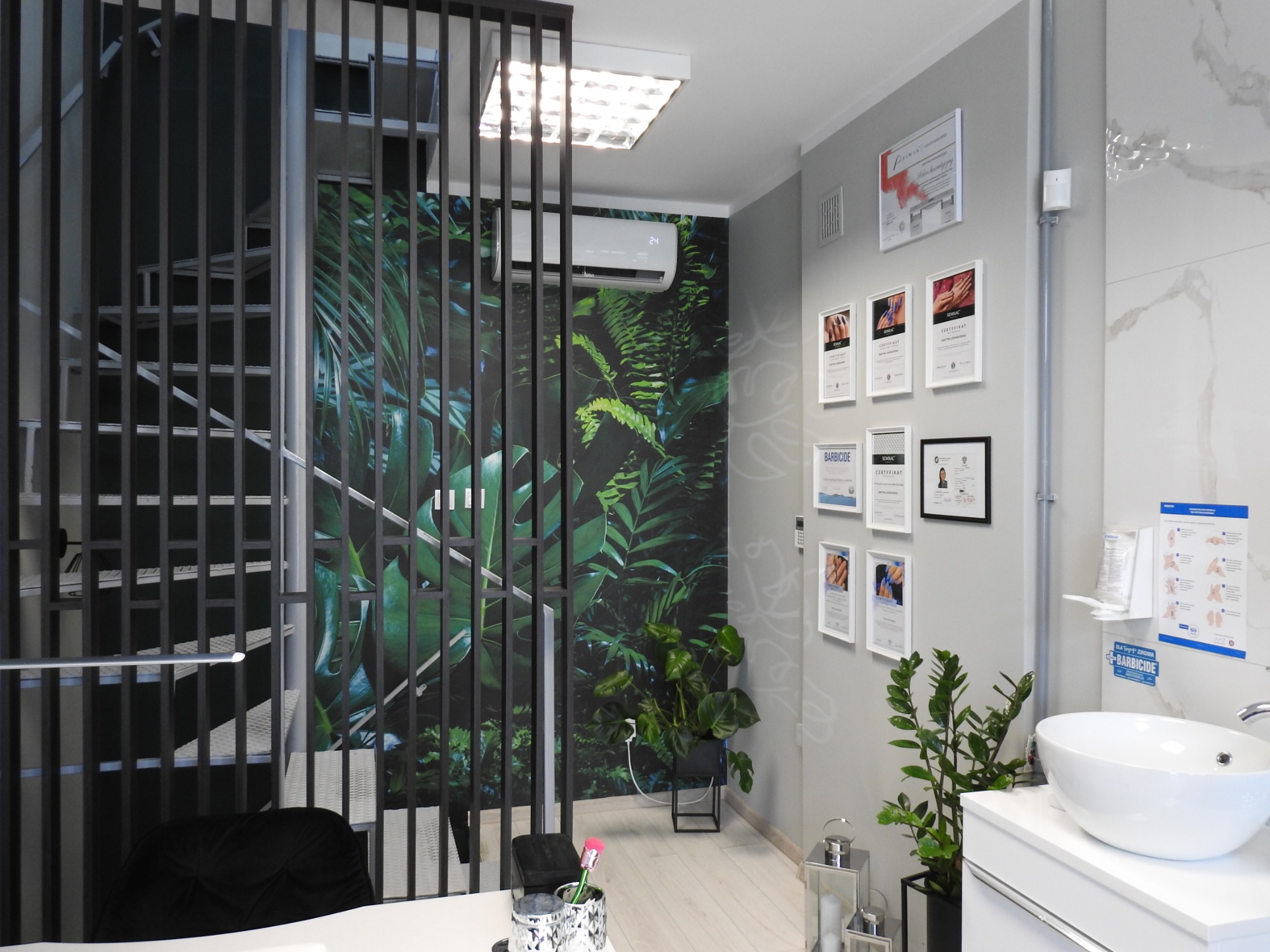 Nowoczesne wnętrze salonu kosmetycznego z motywem tropikalnym na ścianie, oddzielone ażurowym przepierzeniem od schodów, z umywalką na białym blacie i certyfikatami na ścianie.