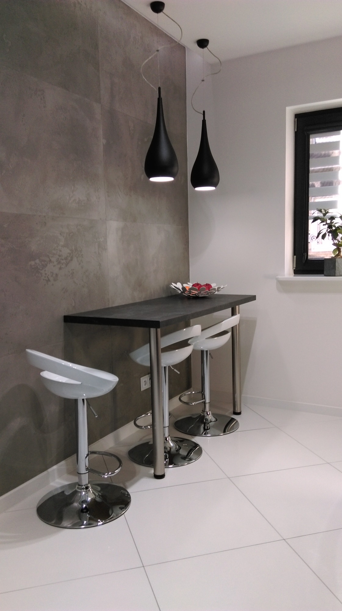 Nowoczesna kuchnia z wysokim stołem barowym, trzema białymi krzesłami z chromowanymi nogami i dwoma czarnymi lampami wiszącymi o kształcie łzy, na ścianie dekoracyjna szara płytka imitująca beton