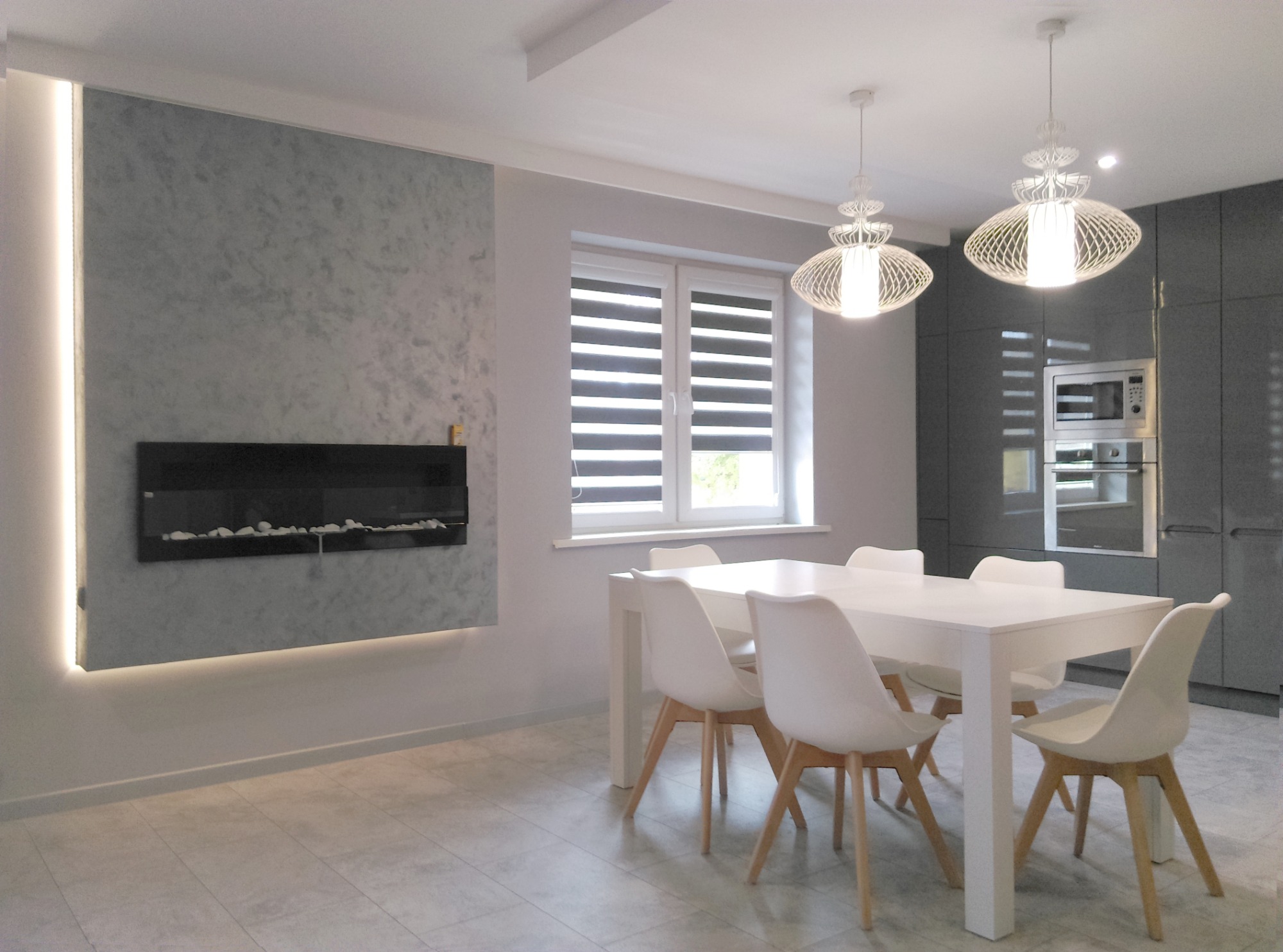 Jasne wnętrze jadalni z białym stołem i krzesłami, dekoracyjnym kominkiem elektrycznym na ścianie z efektem betonu oraz nowoczesnymi lampami wiszącymi.