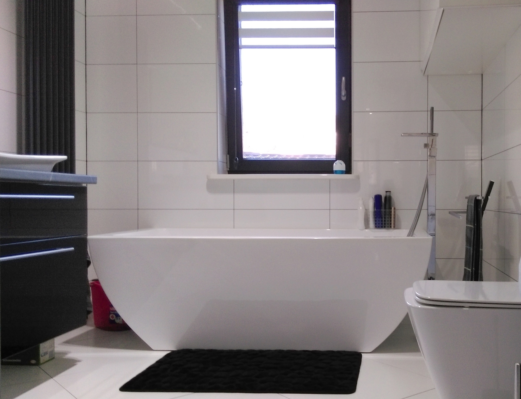 Minimalistyczna łazienka z wolnostojącą wanną, wiszącą toaletą i oknem z roletami dzień-noc, utrzymana w bieli z akcentami czerni.