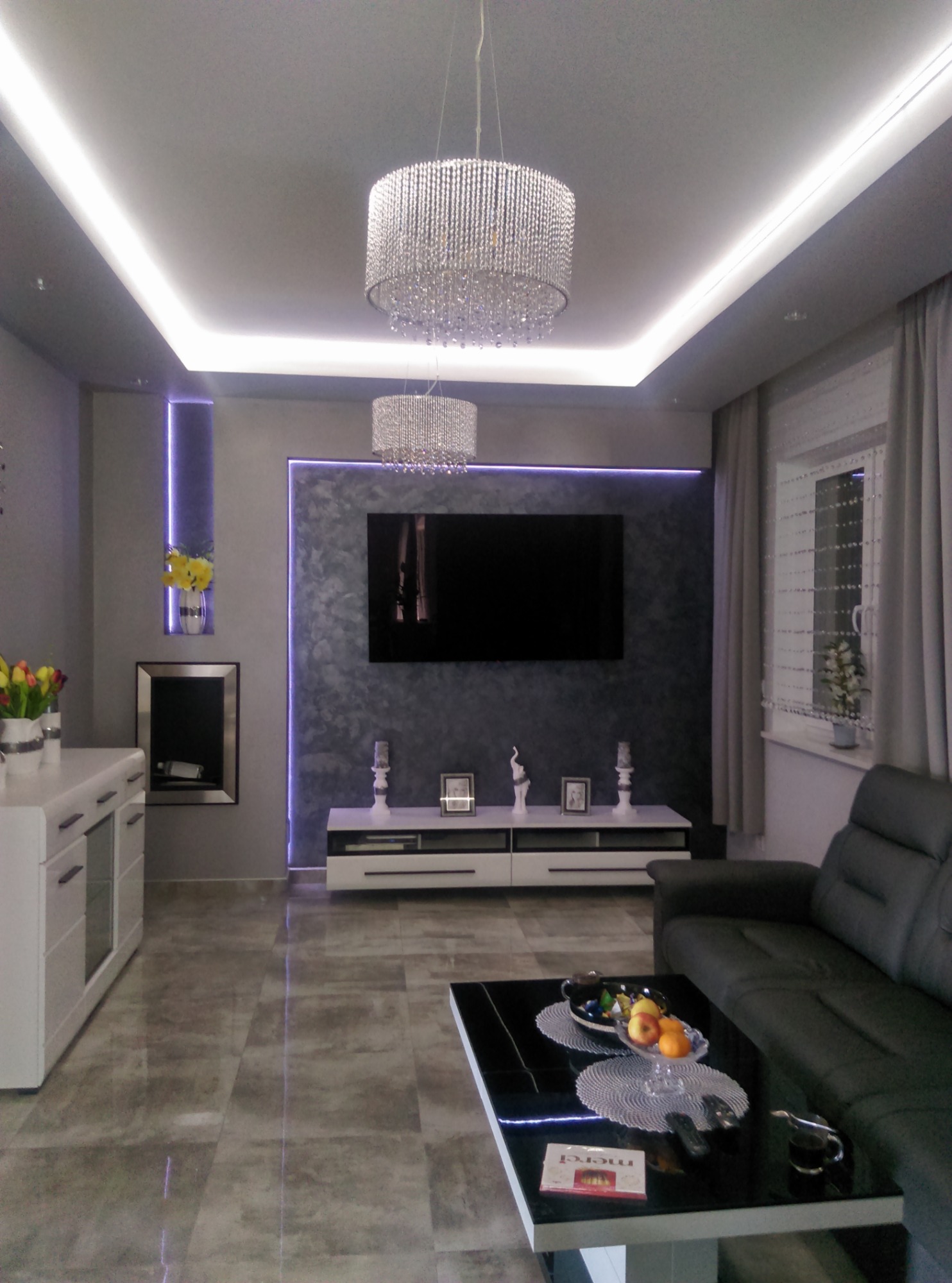 Nowoczesny salon w odcieniach szarości z kryształowymi żyrandolami, podświetleniem LED, telewizorem na ścianie z efektem betonu architektonicznego, białą komodą, czarną sofą i stolikiem kawowym...