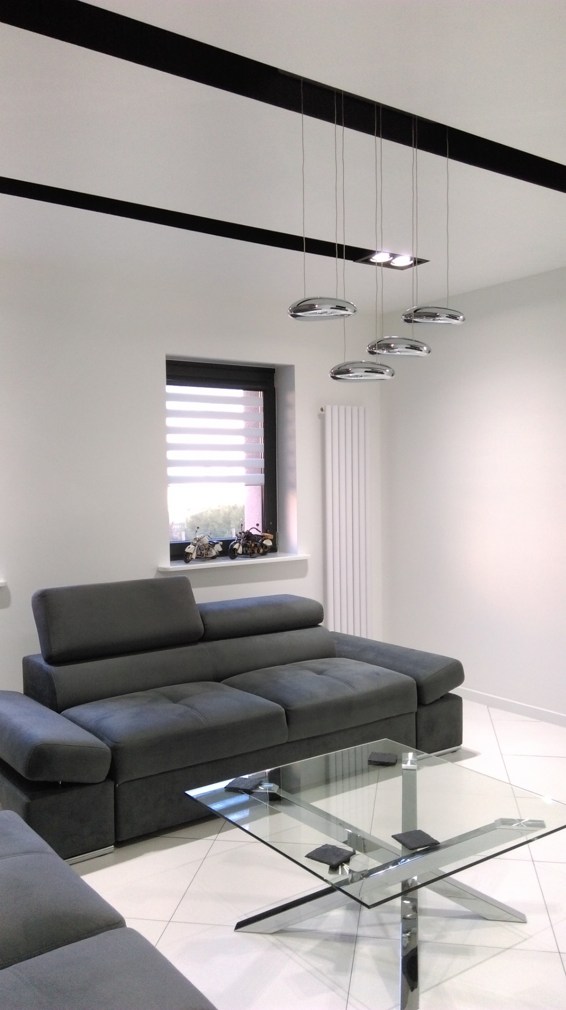 Nowoczesny salon z szarą sofą modułową, szklanym stolikiem kawowym na metalowej podstawie, chromowanymi lampami wiszącymi i dekoracjami w oknie.