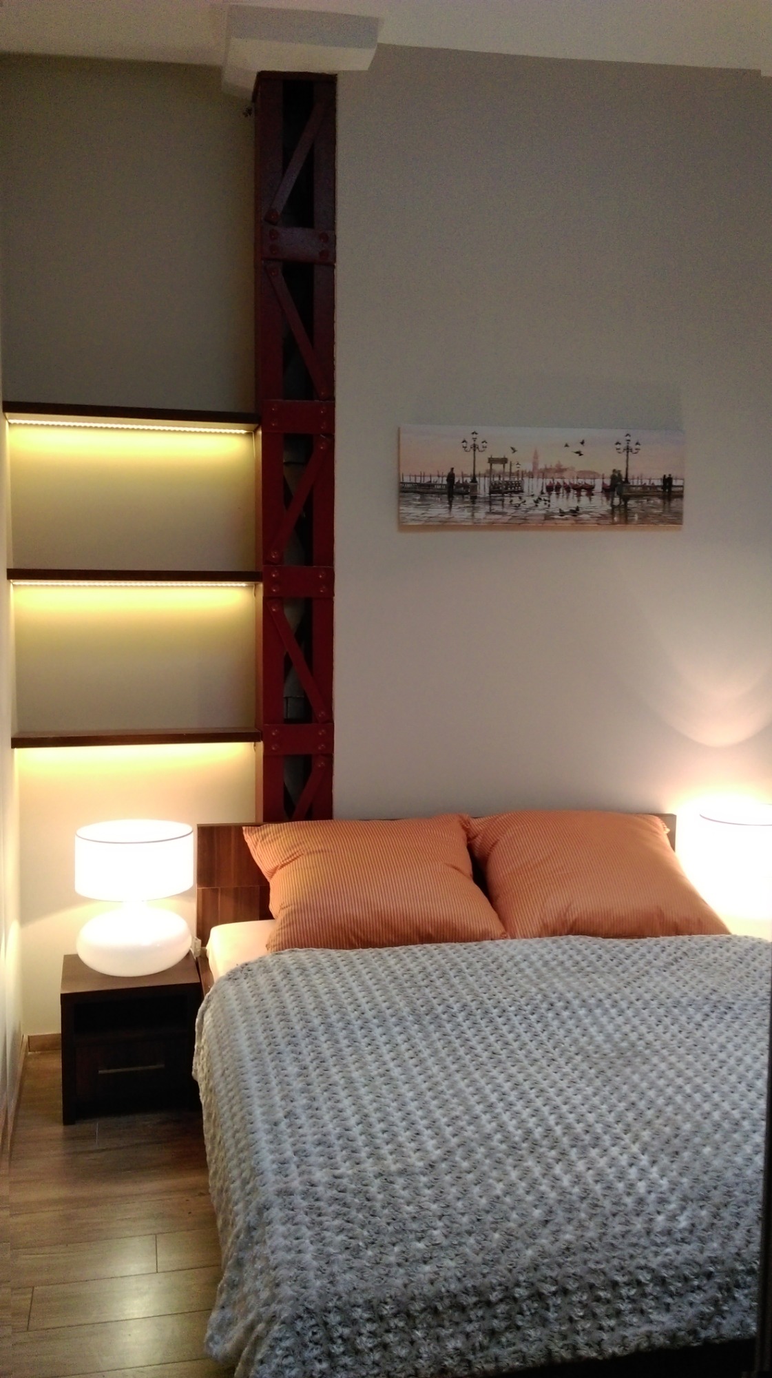 Sypialnia z łóżkiem, pomarańczowymi poduszkami i szarą narzutą, obok szafka nocna z lampką, na ścianie obrazek z motywem weneckim, obok łóżka regał z oświetleniem LED i dekoracyjna, czerwona...