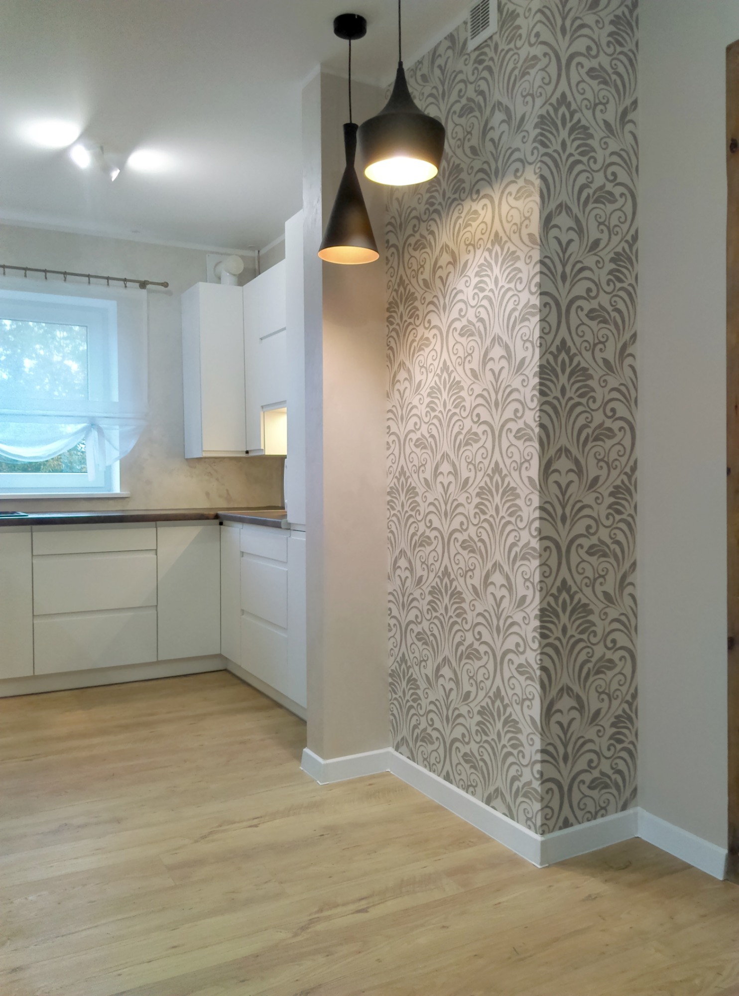 Nowoczesna kuchnia z białymi szafkami, drewnianą podłogą i ozdobną tapetą w odcieniach szarości na filarze, oświetlona dwoma czarnymi lampami wiszącymi.