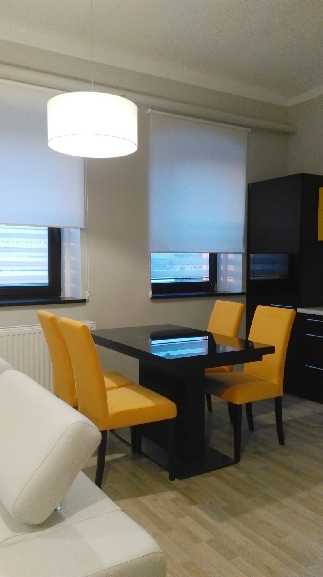 Nowoczesna jadalnia z czarnym stołem i czterema żółtymi krzesłami, oświetlona okrągłą lampą sufitową, z roletami w oknach i fragmentem białej sofy.