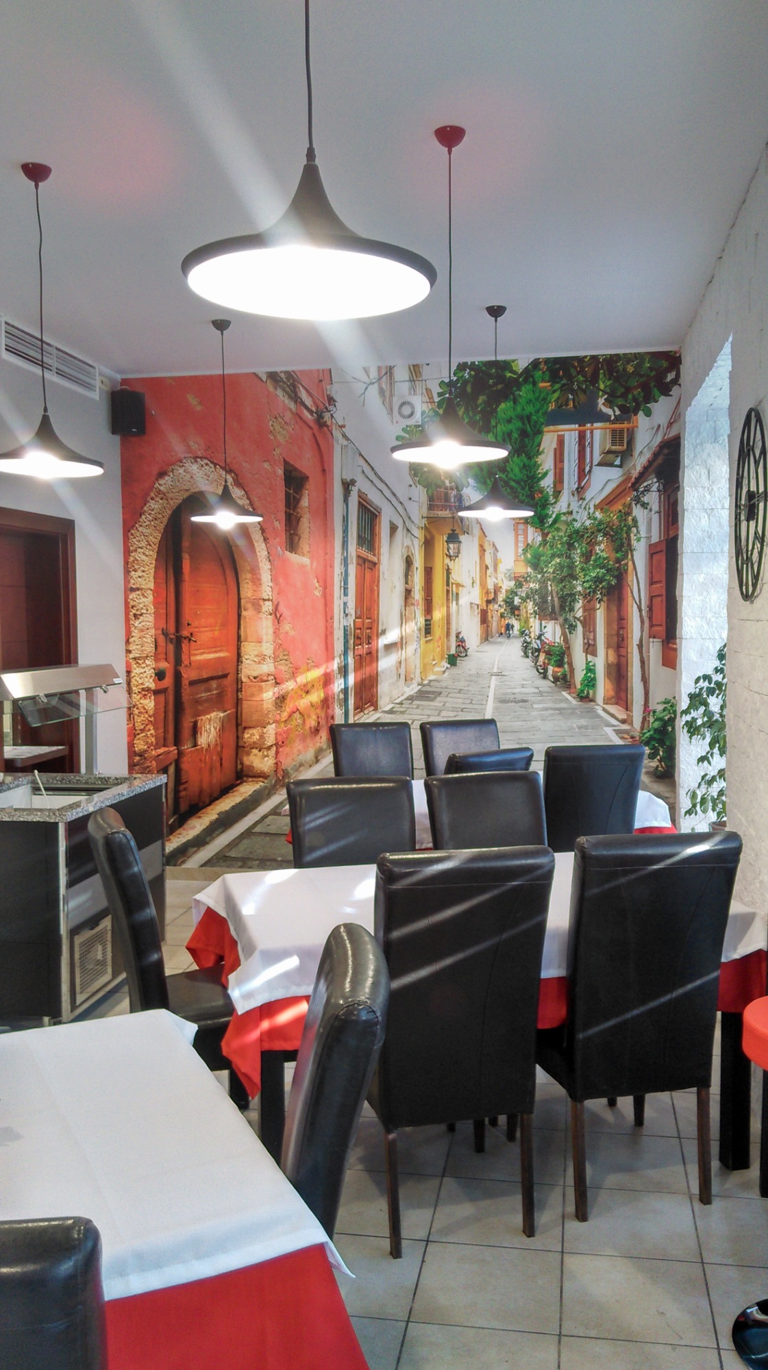 Wnętrze restauracji z fototapetą przedstawiającą uliczkę śródziemnomorskiego miasteczka, czarne krzesła i białe obrusy z czerwonymi akcentami pod okrągłymi lampami.