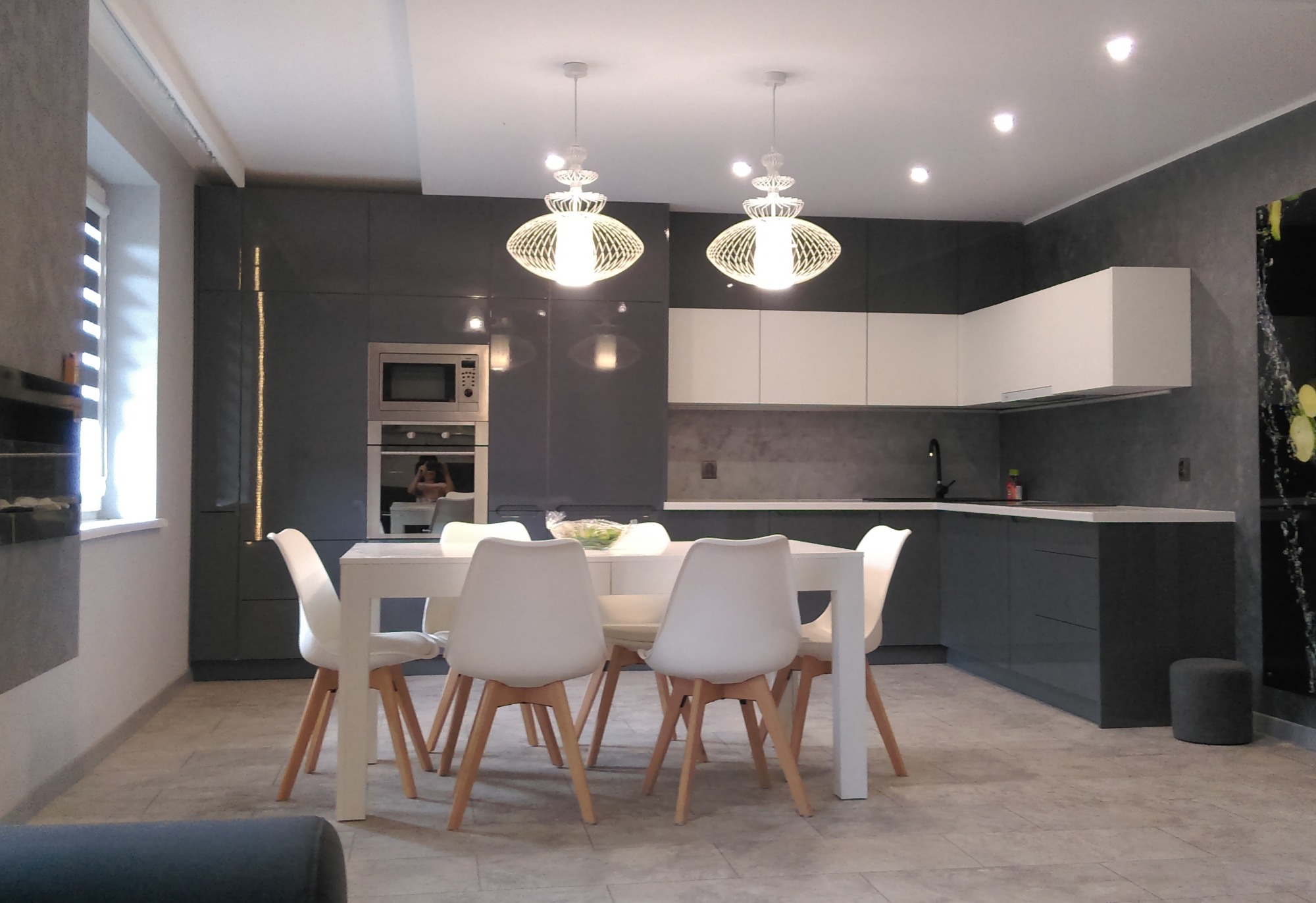 Nowoczesna kuchnia z szarymi szafkami i białymi górnymi szafkami, oświetlona dwoma dekoracyjnymi lampami wiszącymi nad białym stołem z krzesłami.