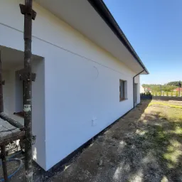 Świeżo otynkowana i pomalowana na biało elewacja domu jednorodzinnego z widocznymi oknami w drewnianych ramach, z fragmentem rusztowania po lewej stronie i niezagospodarowanym terenem wokół budynku.