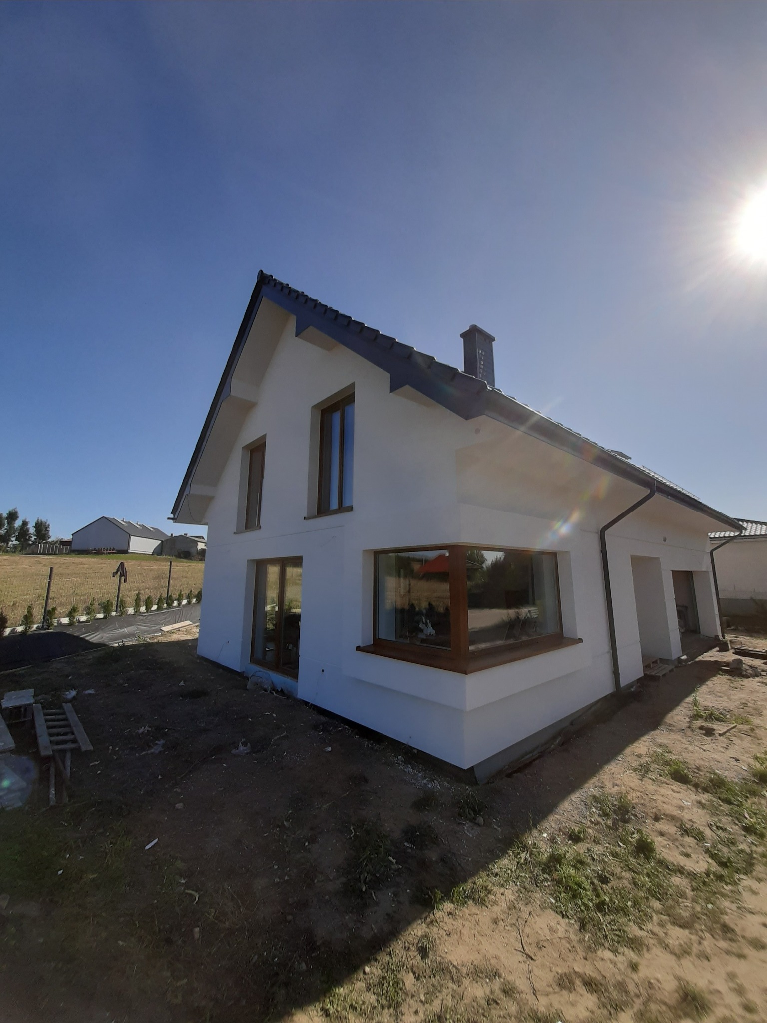 Nowoczesny, biały dom jednorodzinny z ciemną dachówką, świeżo po remoncie elewacji, w słoneczny dzień. Widoczny fragment ogrodu z młodymi tujami.