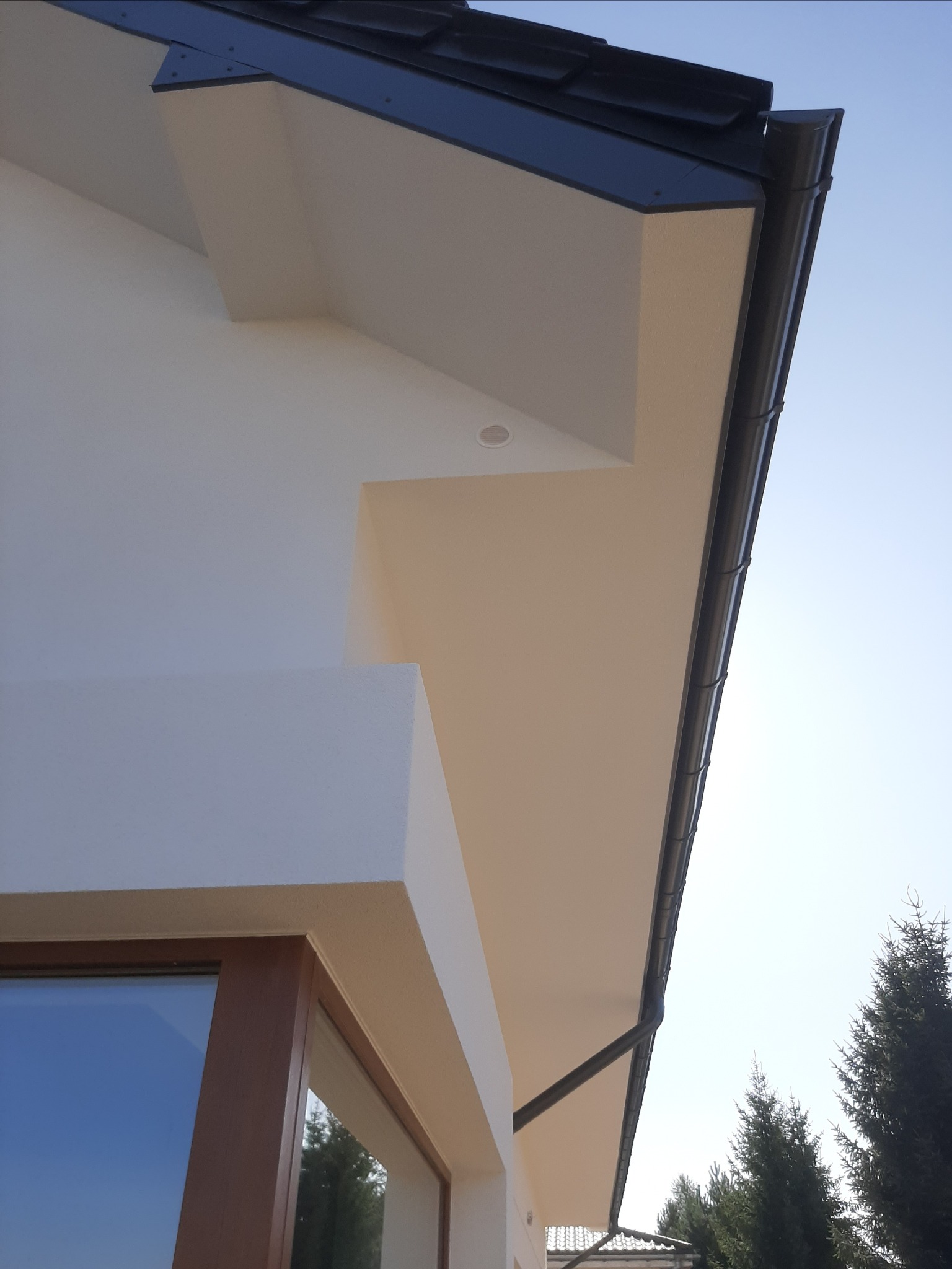 Fragment elewacji domu jednorodzinnego z widocznym oknem, orynnowaniem w kolorze grafitowym i podbitką w odcieniu beżu, oświetlenie punktowe pod dachem.